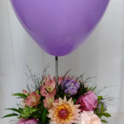 Kompozycja kwiatowa z różowymi, fioletowymi i kremowymi kwiatami w doniczce imitującej korę drzewa, zwieńczona dużym, fioletowym balonem.