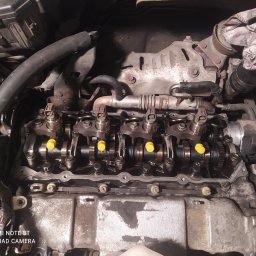 Tomasz Kupis - Otwarty silnik samochodowy z widocznymi wałkami rozrządu, popychaczami i żółtymi zaślepkami w trakcie naprawy lub serwisu.