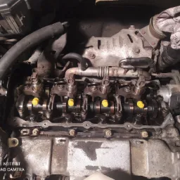 Otwarty silnik samochodowy z widocznymi wałkami rozrządu, popychaczami i żółtymi zaślepkami w trakcie naprawy lub serwisu.