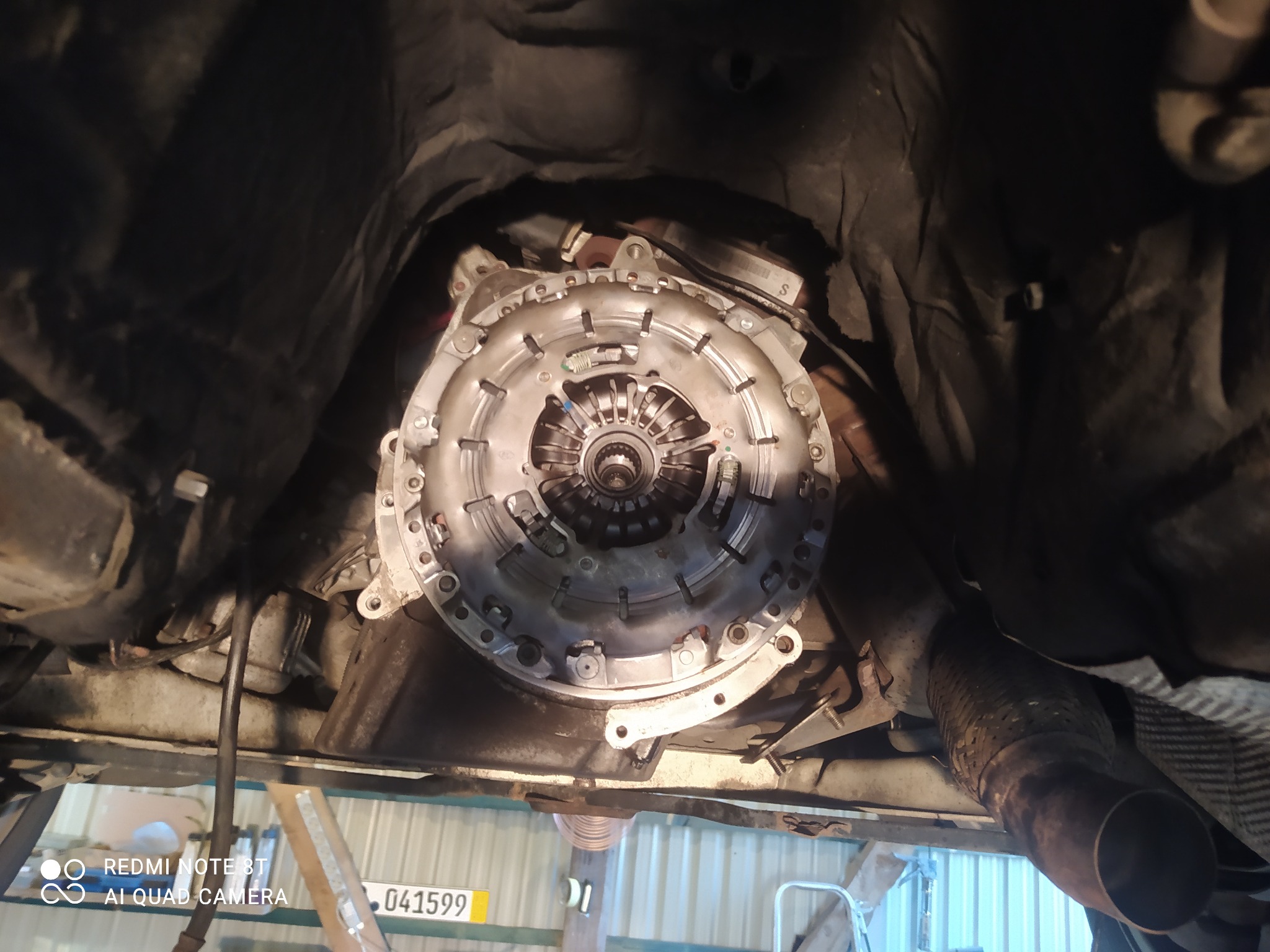 Widok od spodu na sprzęgło samochodowe zamontowane w pojeździe podczas naprawy, widoczny fragment układu wydechowego i podnośnik warsztatowy.