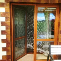 Drewniane okno z moskitierą na zawiasach, częściowo otwarte, z widokiem na ogród przez firankę w drugim oknie, na zewnątrz dywanik z motywem ulicy.