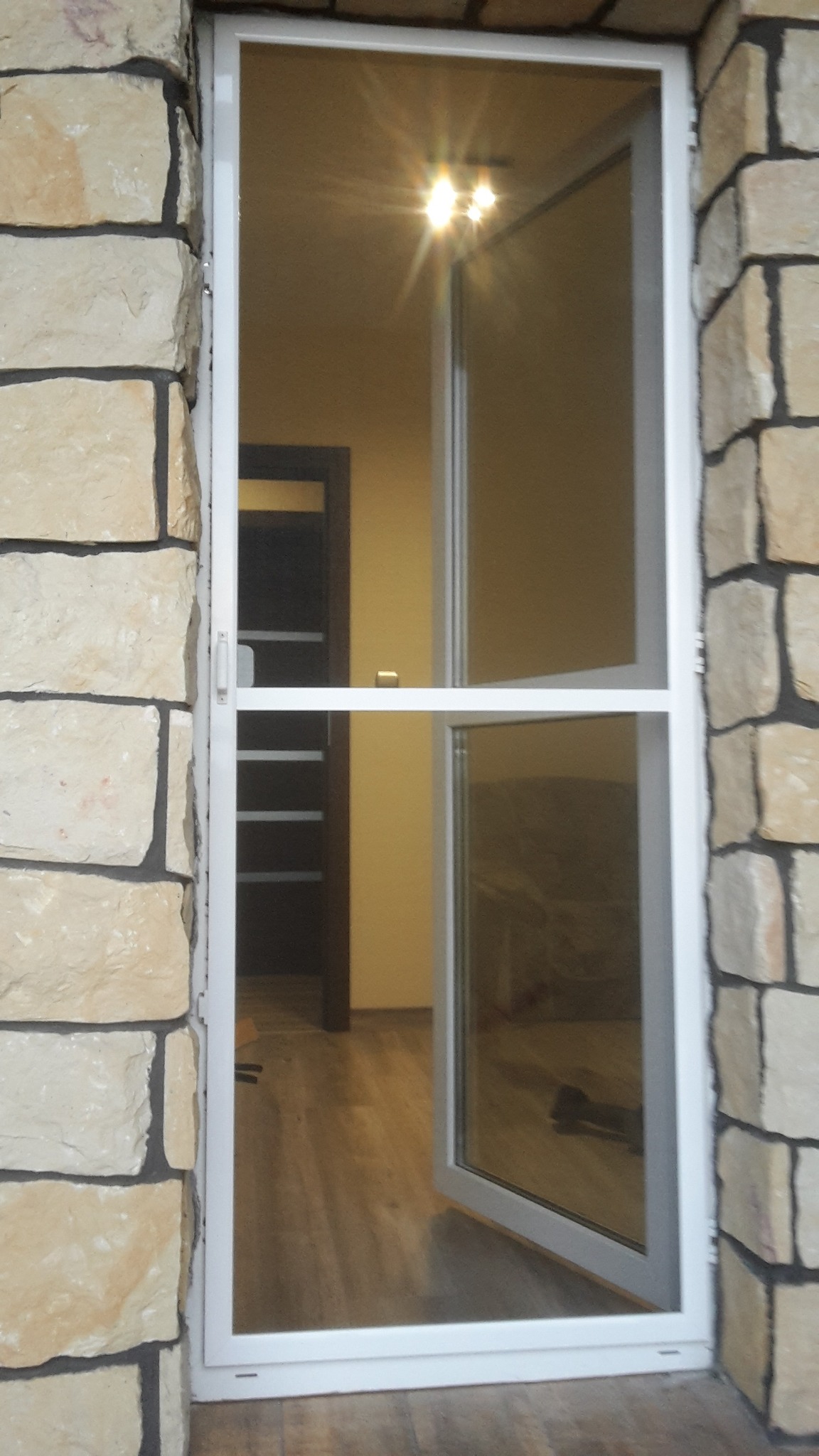 Otwarta, biała rama z siatką w oknie, zamontowana w kamiennej ścianie; widoczny fragment wnętrza z drewnianą podłogą i drzwiami.