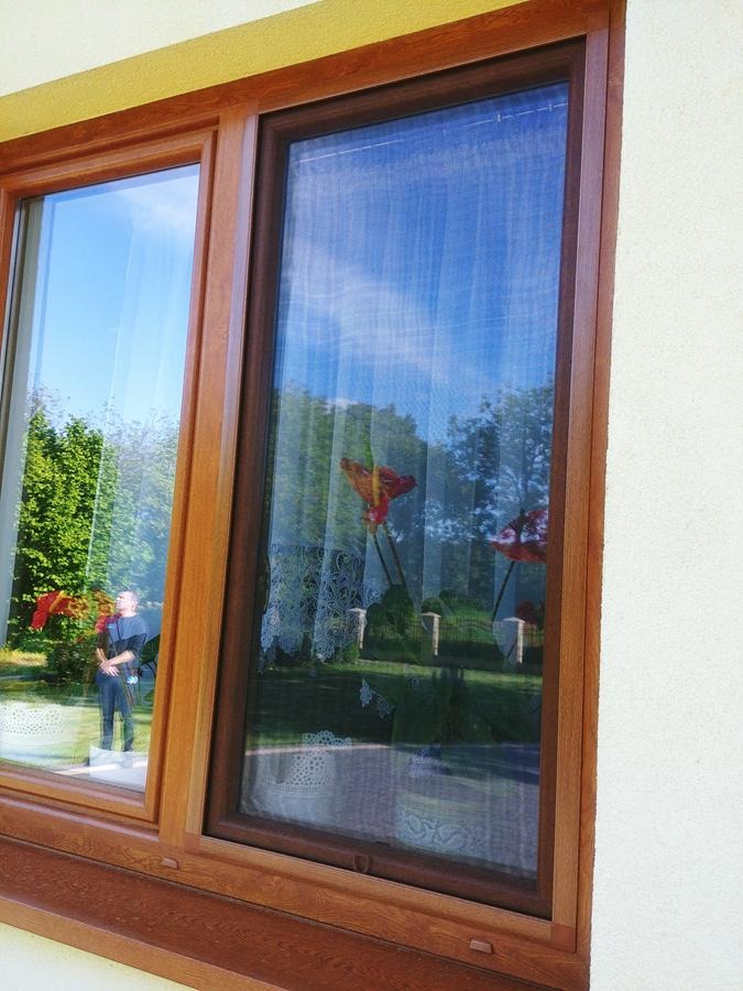 Drewniane okno z zamontowaną siatką ochronną przed owadami, widok z zewnątrz, Stopnica; w szybie okna odbija się osoba i zieleń ogrodu.