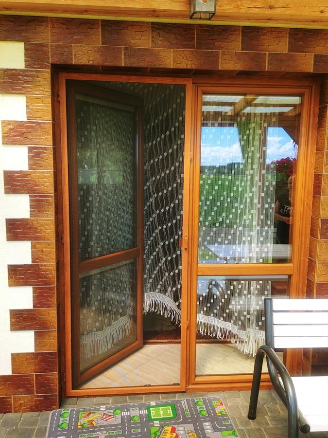 Drewniane okno z moskitierą na zawiasach, częściowo otwarte, z widokiem na ogród przez firankę w drugim oknie, na zewnątrz dywanik z motywem ulicy.