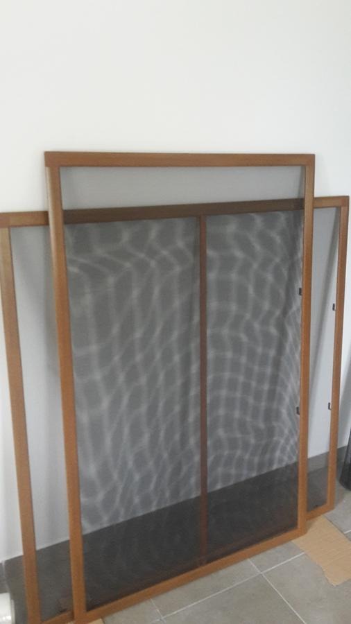 Dwie moskitiery z brązowymi ramkami, jedna oparta o ścianę, druga częściowo widoczna za nią, na podłodze w jasnym pomieszczeniu.