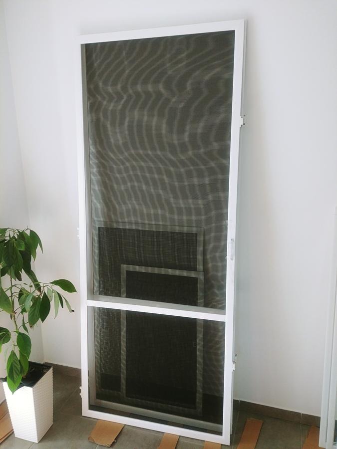 Biała rama z siatką przeciw owadom, widoczne trzy segmenty siatki o różnych rozmiarach, obok doniczka z zieloną rośliną.