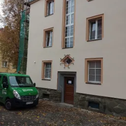 Odnowiona elewacja budynku z beżową farbą i brązowymi obramowaniami okien, widoczny fragment rusztowania z zieloną siatką, firmowy zielony van z logo TG System na tle jesiennych liści.