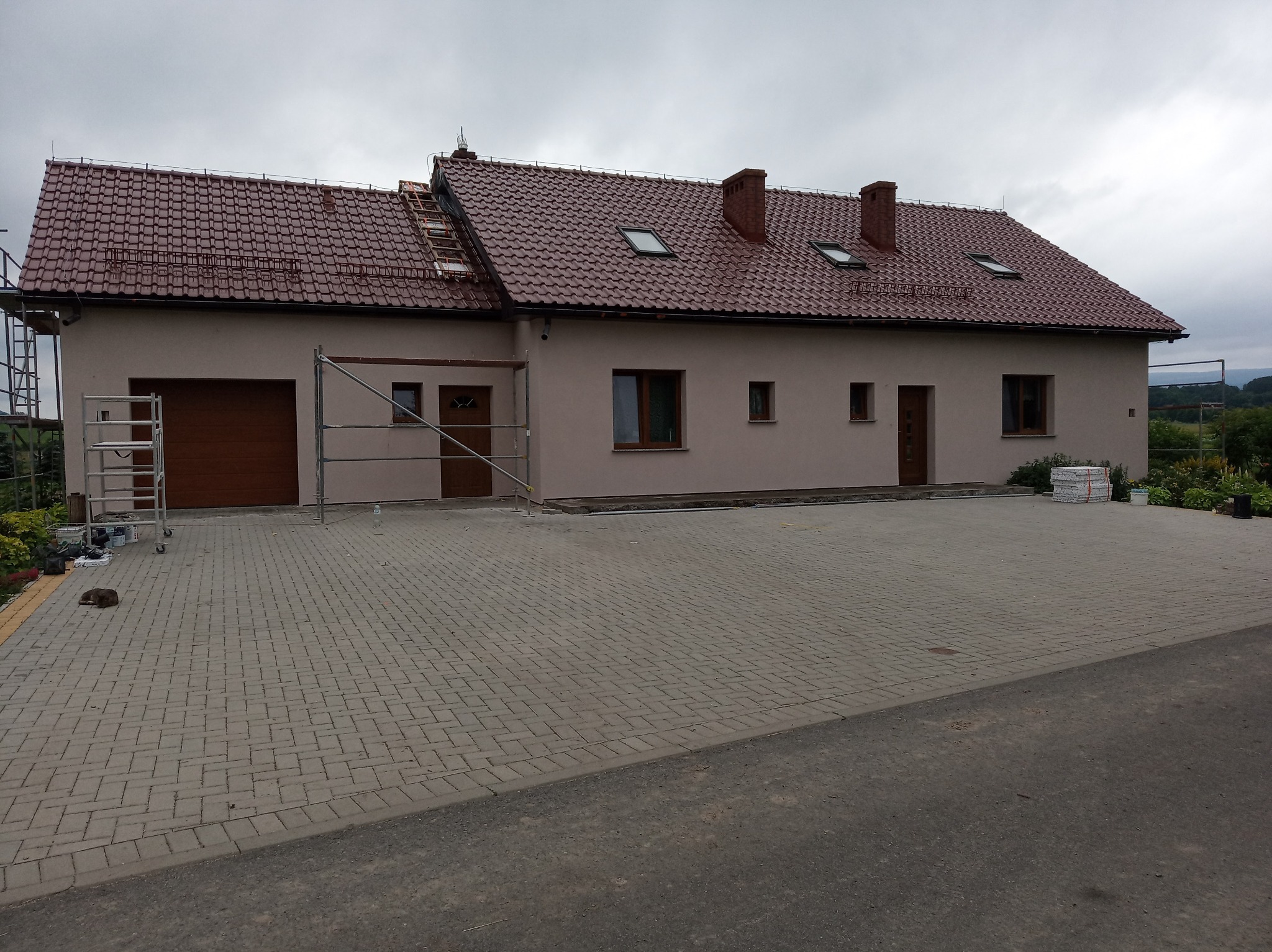 Dwukondygnacyjny dom z beżową elewacją, czerwoną dachówką i brązowymi oknami, częściowo otoczony rusztowaniem, z ułożoną kostką brukową na podjeździe i leżącym kotem.