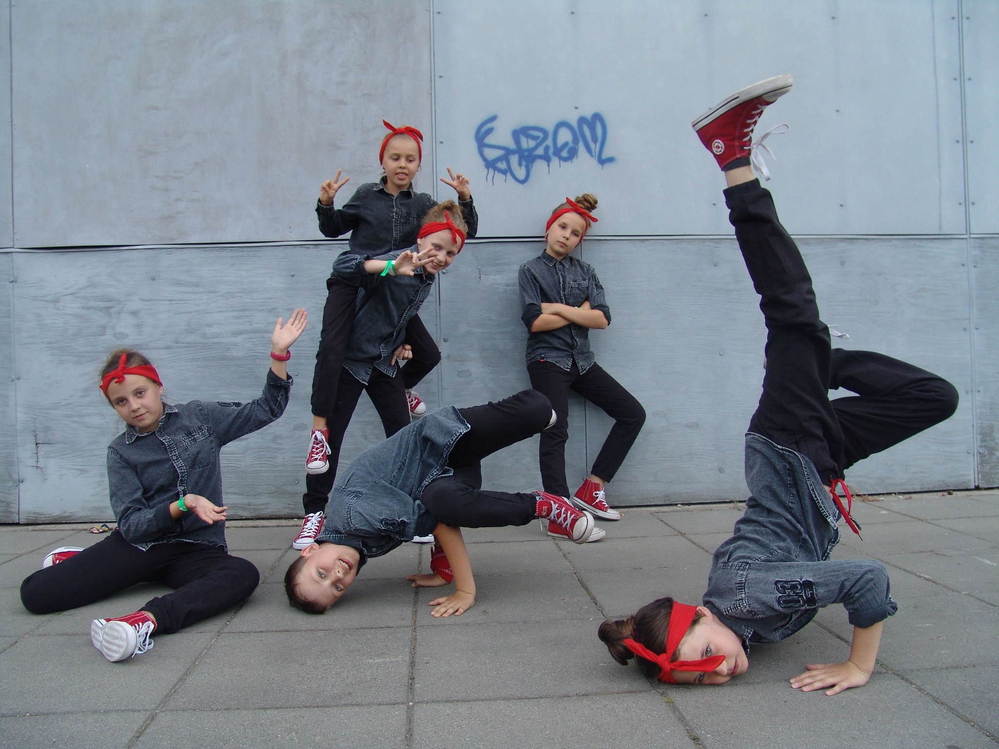 Grupa dzieci w czerwonych bandanach i dżinsowych koszulach prezentuje układy taneczne na tle szarej ściany z graffiti.