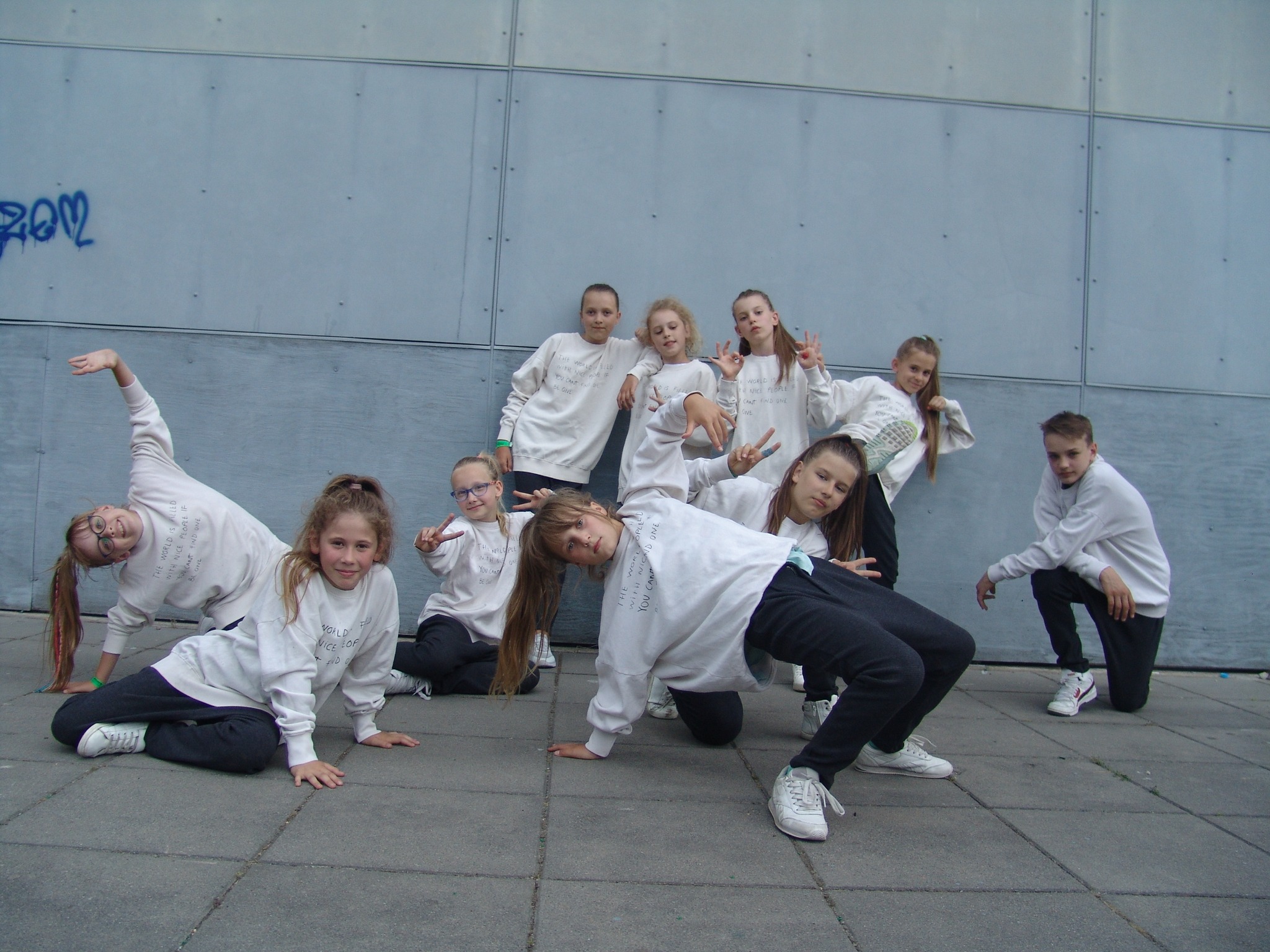 Grupa dzieci w białych bluzach z napisami pozuje do zdjęcia na tle szarej ściany z graffiti, wykonując figury taneczne.