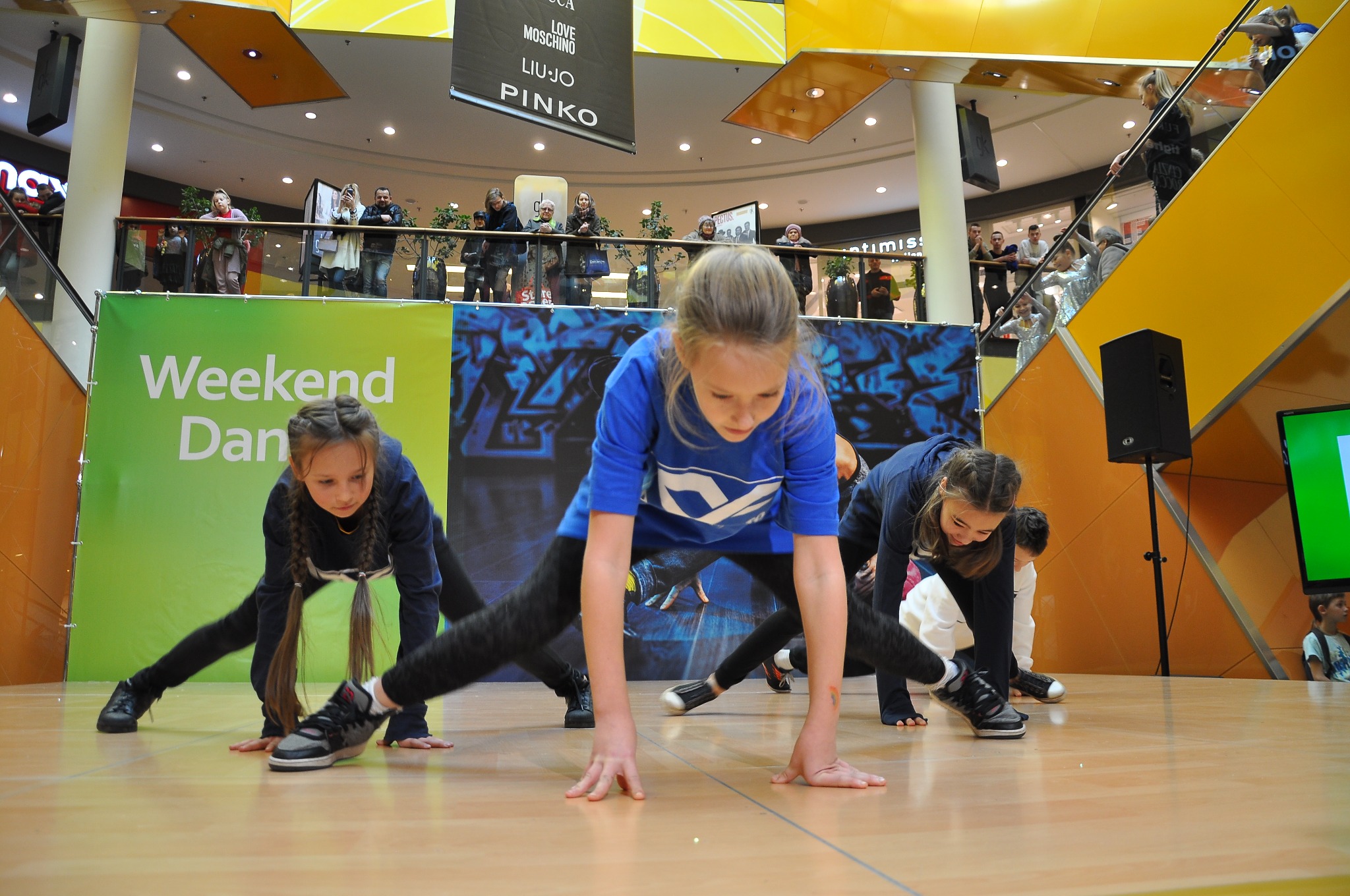 Grupa młodych tancerzy w rozkroku na scenie podczas występu w centrum handlowym, widzowie obserwują z góry.