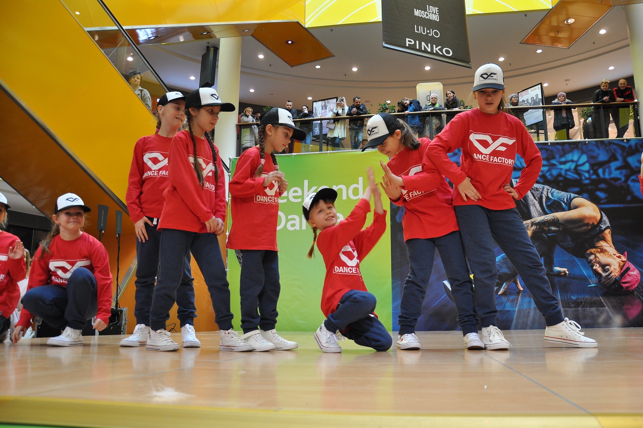 Grupa dzieci w czerwonych koszulkach z logo Dance Factory prezentuje układ taneczny na scenie w centrum handlowym, obserwowane przez publiczność na balkonie