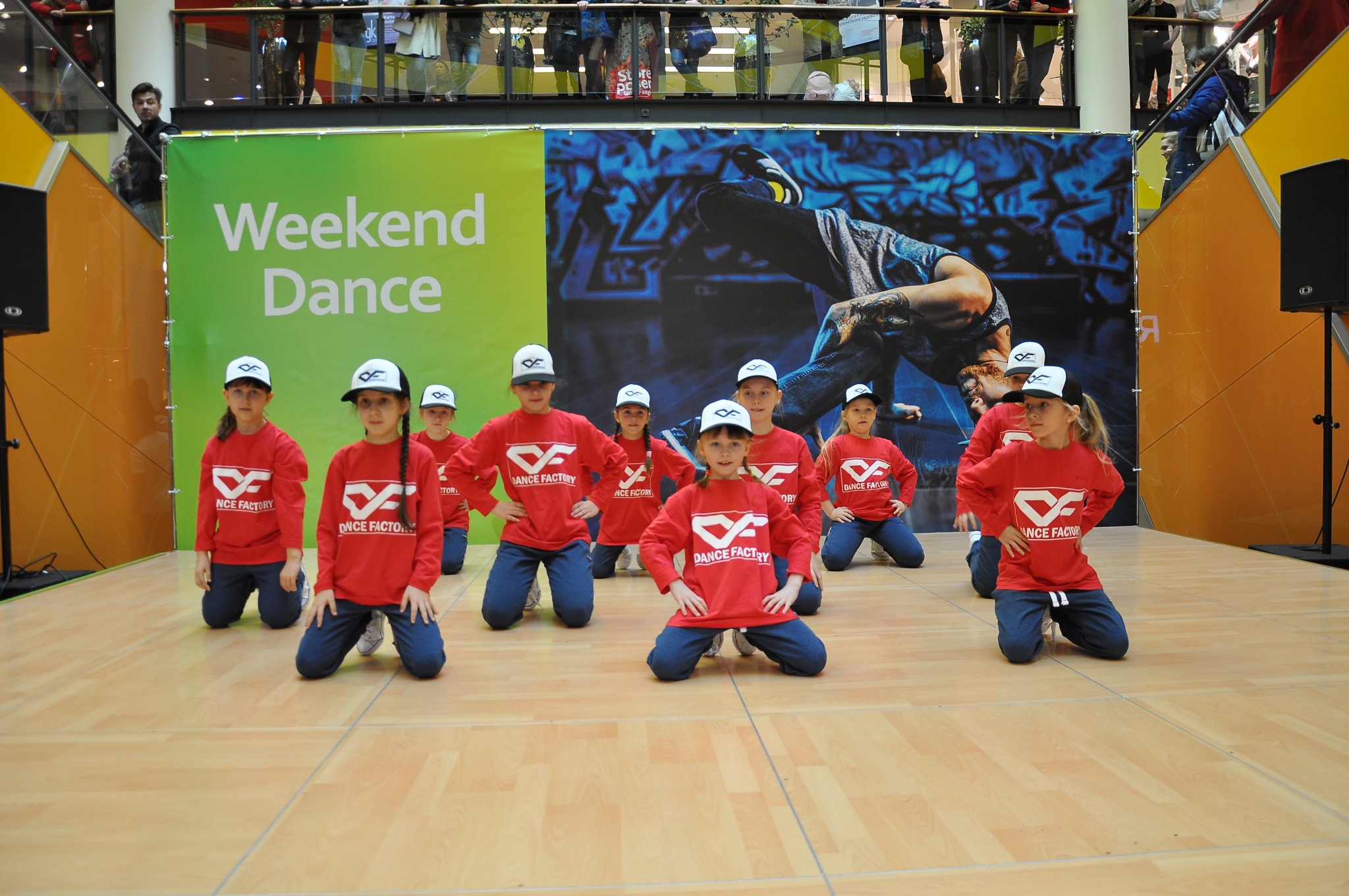 Grupa dzieci w czerwonych koszulkach z logo 'Dance Factory' i czapkach, klęczących na scenie podczas występu 'Weekend Dance' w galerii handlowej.