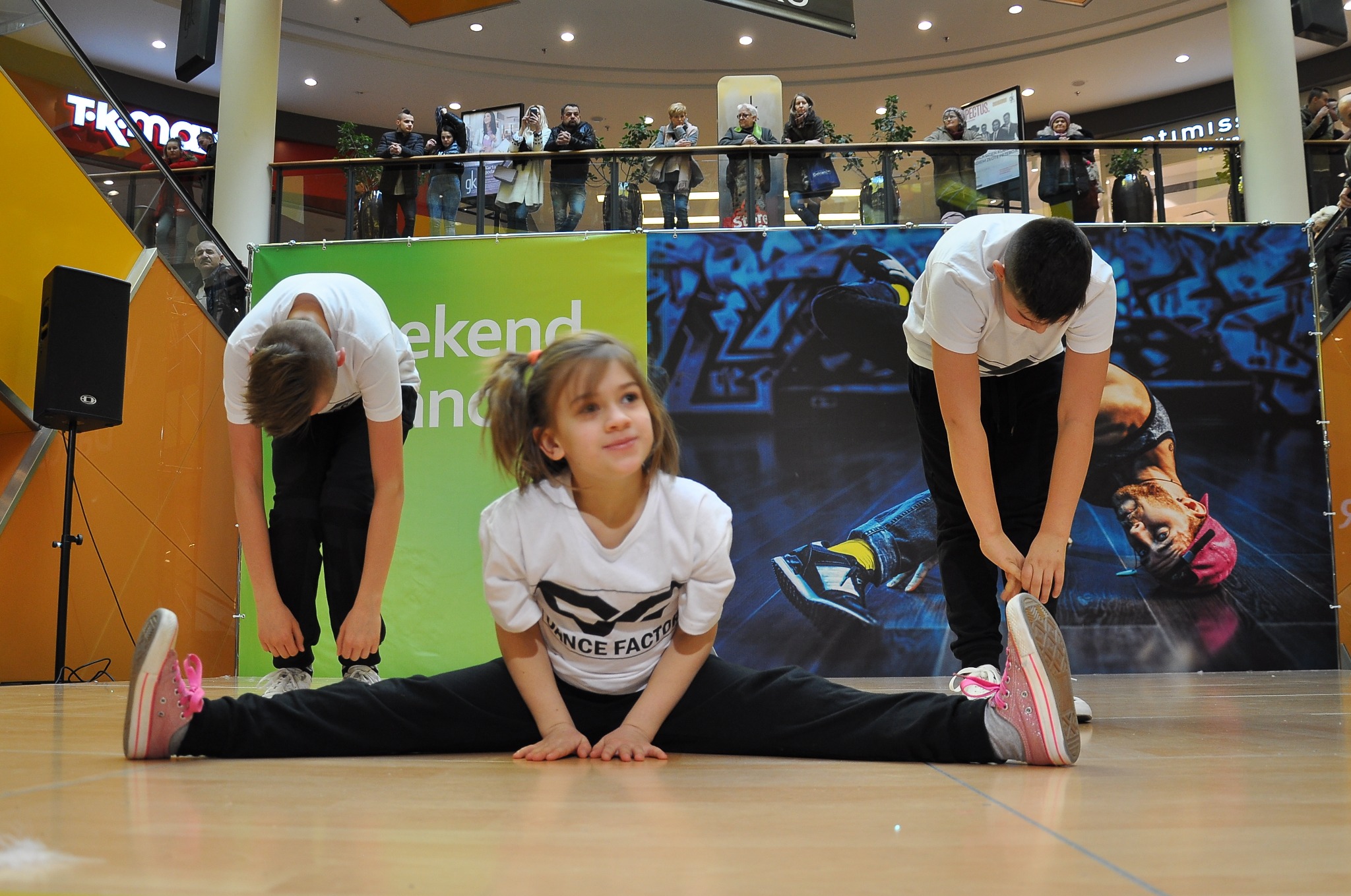 Młoda dziewczyna w rozkroku na scenie, ubrana w białą koszulkę z napisem 'DANCE FACTOR', obok dwóch nastolatków w białych koszulkach, rozciągających się, w tle plakat z tancerzem i publiczność...