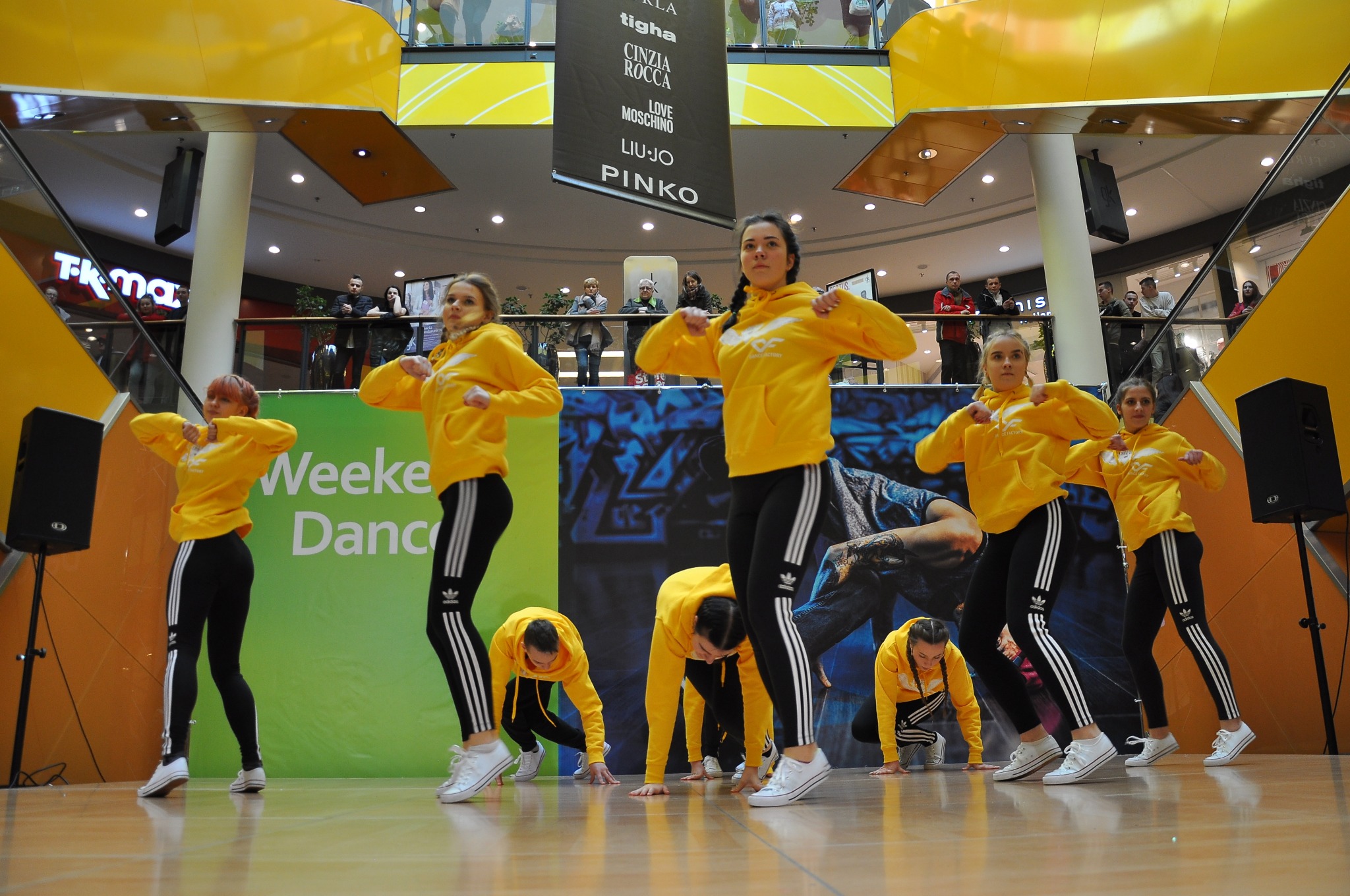 Grupa tancerzy w żółtych bluzach z kapturem i czarnych legginsach w trakcie występu tanecznego na scenie w centrum handlowym.