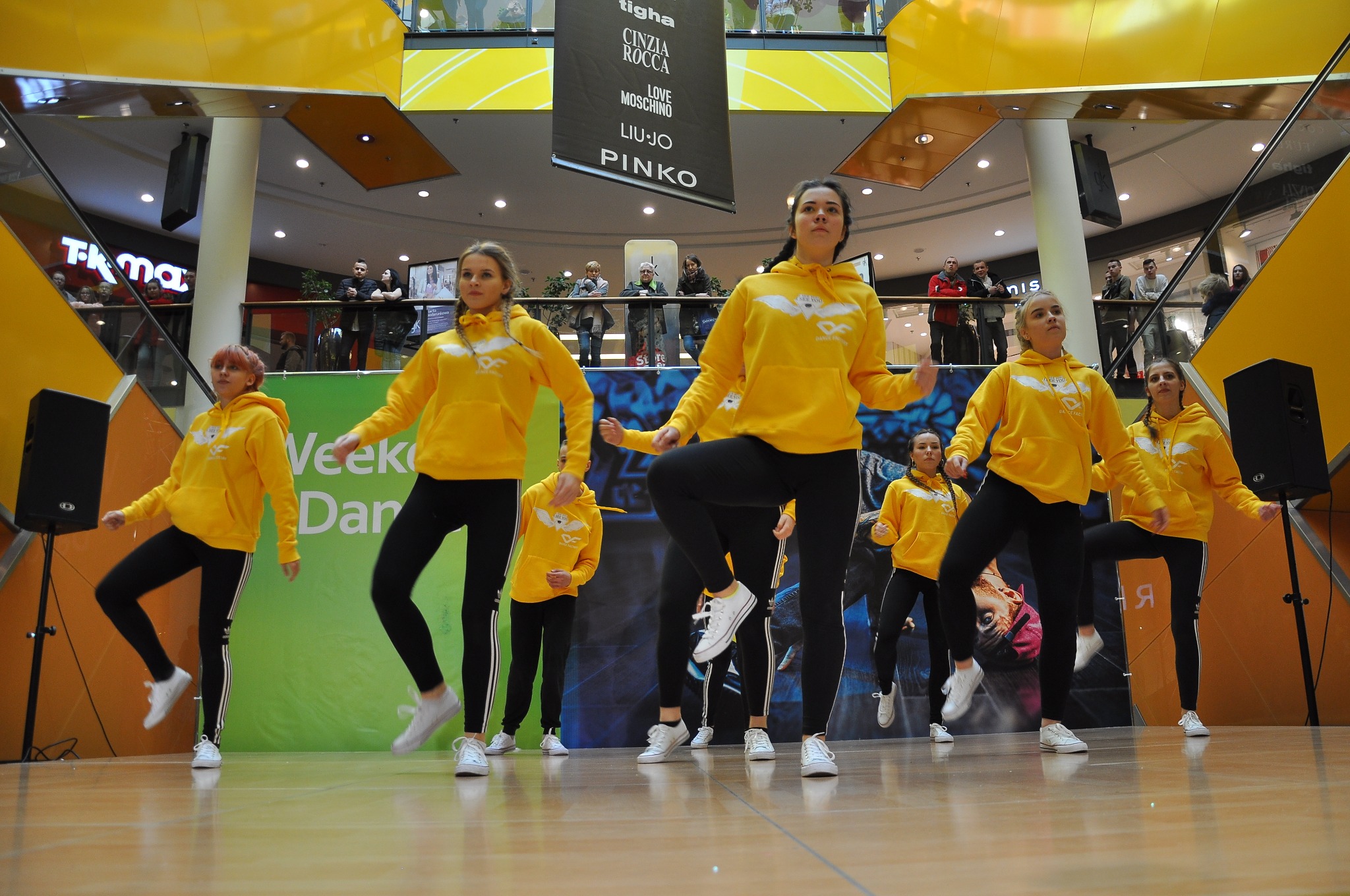 Grupa tancerek w żółtych bluzach z logo prezentuje choreografię na scenie w centrum handlowym, widoczni widzowie na wyższych poziomach.