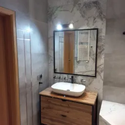 Nowoczesna łazienka z umywalką nablatową na drewnianej szafce, lustro w czarnej ramie z oświetleniem, marmurowe płytki na ścianie i grzejnik drabinkowy widoczny w odbiciu.