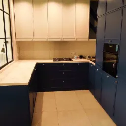 Nowoczesna kuchnia w odcieniach granatu i bieli z marmurowym blatem, widok na płytę grzewczą i okno z czarnymi szprosami, podłoga wyłożona płytkami imitującymi drewno.