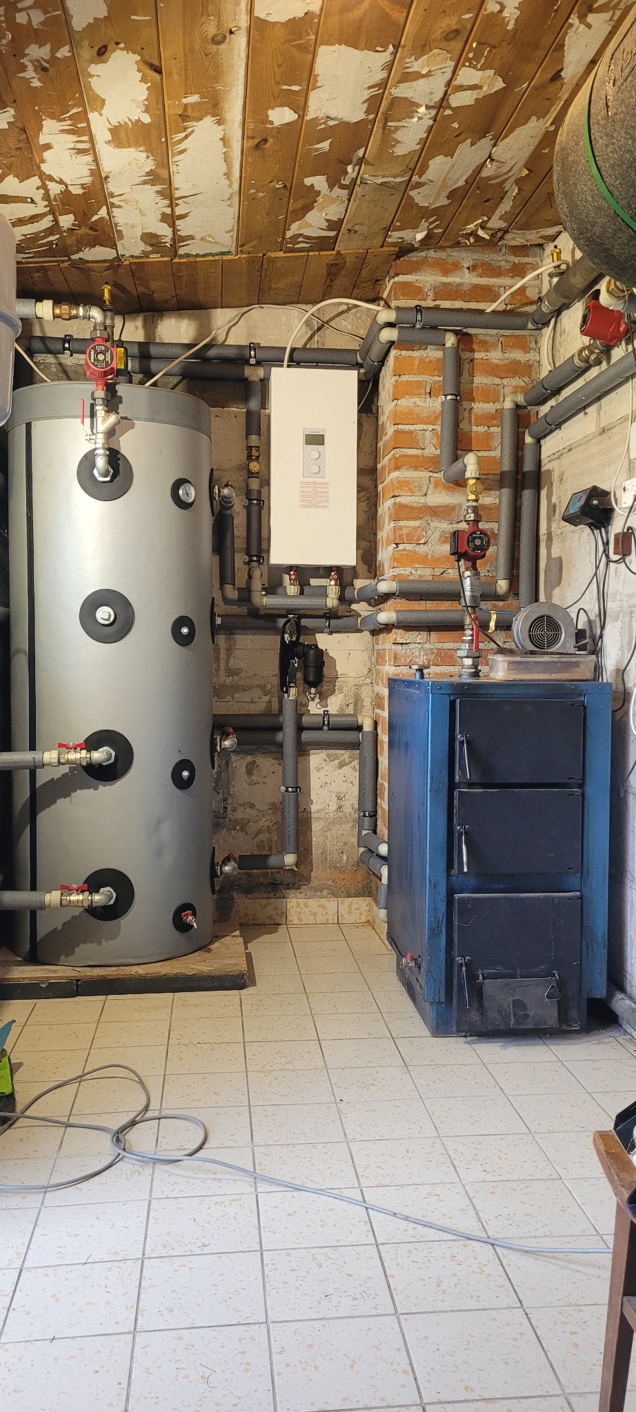 Kotłownia z piecem na paliwo stałe, zasobnikiem ciepłej wody i instalacją grzewczą. Widoczne rury, zawory i sterownik. Ściany z cegły i drewniana boazeria.