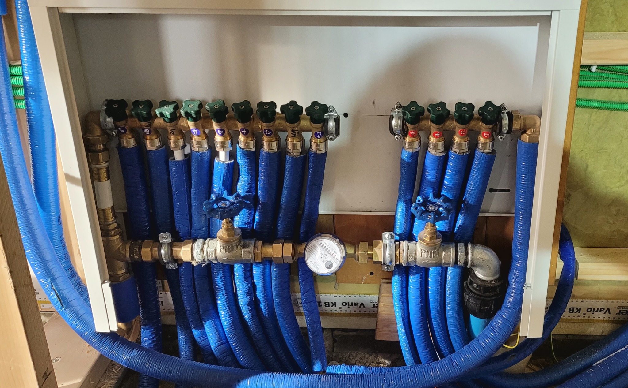 Rozdzielacz instalacji wodnej z licznymi niebieskimi rurami, zaworami z zielonymi pokrętłami i wodomierzem, umieszczony w białej obudowie w trakcie montażu.