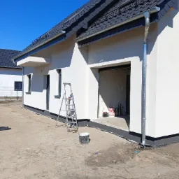 Nowo otynkowana, biała elewacja domu jednorodzinnego z szarą podmurówką, widoczna drabina oparta o ścianę, puste wiadro i puszki po farbie w otwartym garażu.