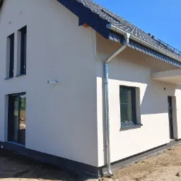 Nowoczesny dom jednorodzinny z jasną elewacją, ciemną stolarką okienną i dachem z dachówki ceramicznej, widoczny system rynnowy i przygotowany teren wokół budynku.