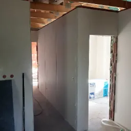 Wnętrze w trakcie prac wykończeniowych: zamontowane płyty kartonowo-gipsowe tworzą korytarz, widoczne drewniane belki stropowe, izolacja wokół ościeżnicy i porozrzucane resztki materiałów...