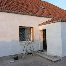 Dom z częściowo pomalowaną elewacją na jasnoszaro, drabina oparta o ścianę przy oknie, wiadro z farbą obok, dach z czerwonej dach&oacute;wki.