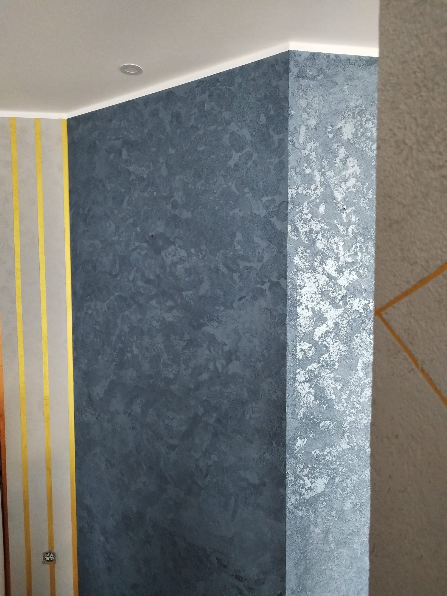 Ściana wewnętrzna z dekoracyjnym tynkiem w odcieniach szarości i błękitu, pionowe złote pasy na sąsiedniej ścianie, oświetlenie punktowe sufitowe.