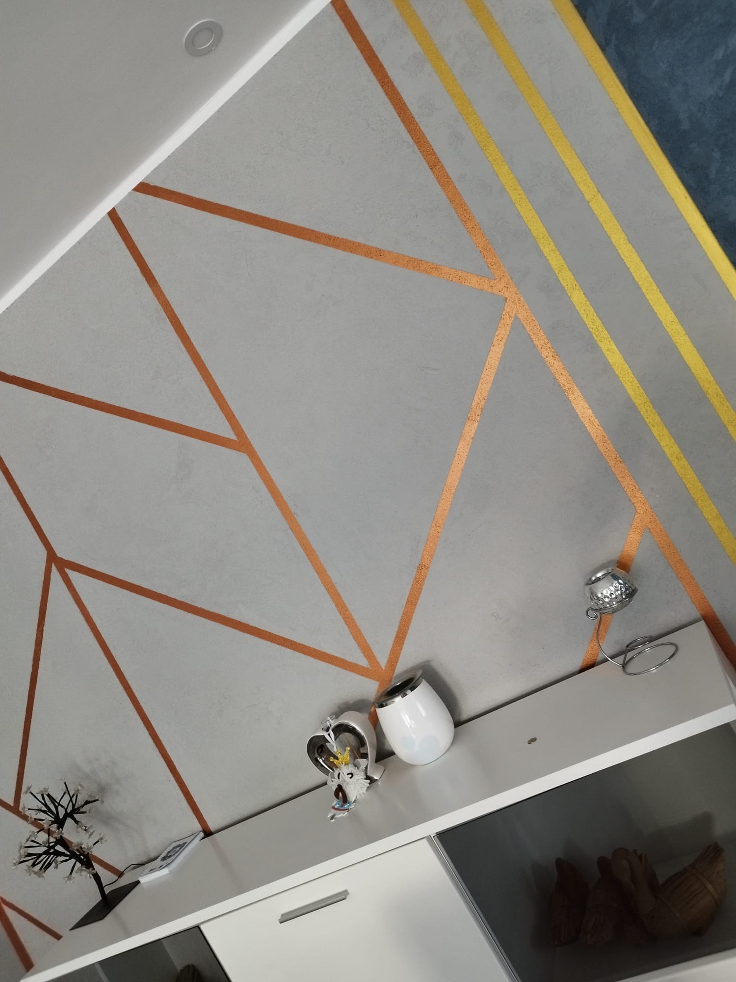 Dekoracyjny sufit z szarego tynku strukturalnego z geometrycznymi wzorami w kolorze miedzianym i złotym, oświetlenie punktowe i ozdobne przedmioty na szafce pod sufitem.