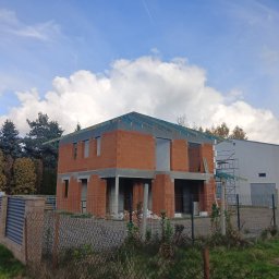 Krystian Wasilewski Usługi Remontowo-Budowlane - Dwukondygnacyjny dom w stanie surowym otwartym, z czerwonej cegły, z widoczną konstrukcją dachu i elementami szalunków, ogrodzony siatką, na tle błękitnego nieba z chmurami i zieleni