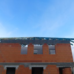 Krystian Wasilewski Usługi Remontowo-Budowlane - Wznoszenie dwukondygnacyjnego domu z czerwonej cegły ceramicznej, widoczne otwory okienne i fragment drewnianej konstrukcji dachu na tle błękitnego nieba.