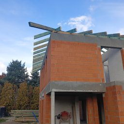 Krystian Wasilewski Usługi Remontowo-Budowlane - Surowy stan domu murowanego z czerwonej cegły ceramicznej z widocznymi elementami szalunku i drewnianymi belkami więźby dachowej na tle błękitnego nieba z chmurami
