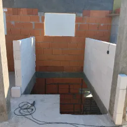 Wznoszenie ścian fundamentowych z bloczków. Widoczne zbrojenie i otwór okienny w murze z czerwonej cegły. Na pierwszym planie narzędzie do cięcia.