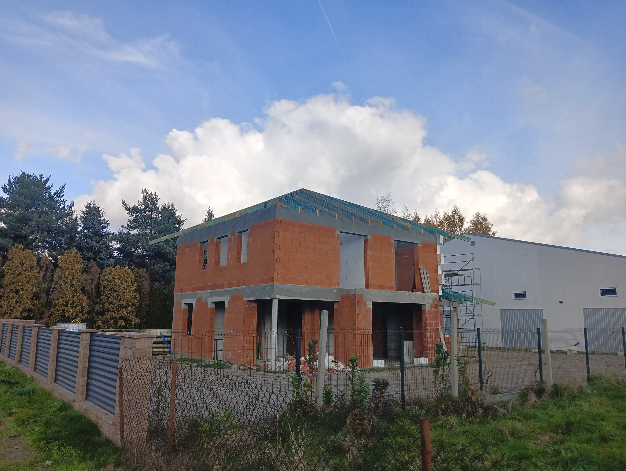 Dwukondygnacyjny dom w stanie surowym otwartym, z czerwonej cegły, z widoczną konstrukcją dachu i elementami szalunków, ogrodzony siatką, na tle błękitnego nieba z chmurami i zieleni