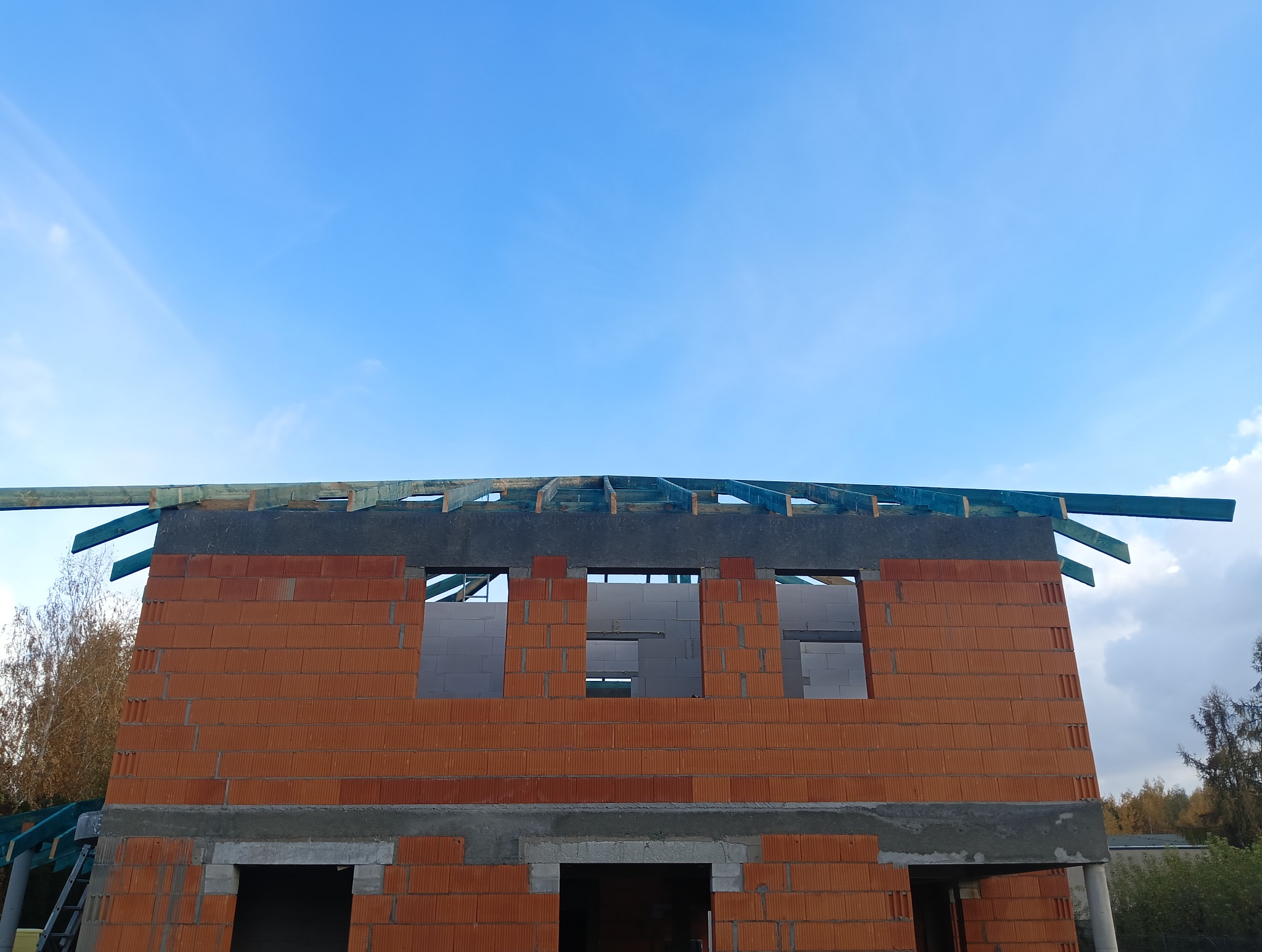 Wznoszenie dwukondygnacyjnego domu z czerwonej cegły ceramicznej, widoczne otwory okienne i fragment drewnianej konstrukcji dachu na tle błękitnego nieba.