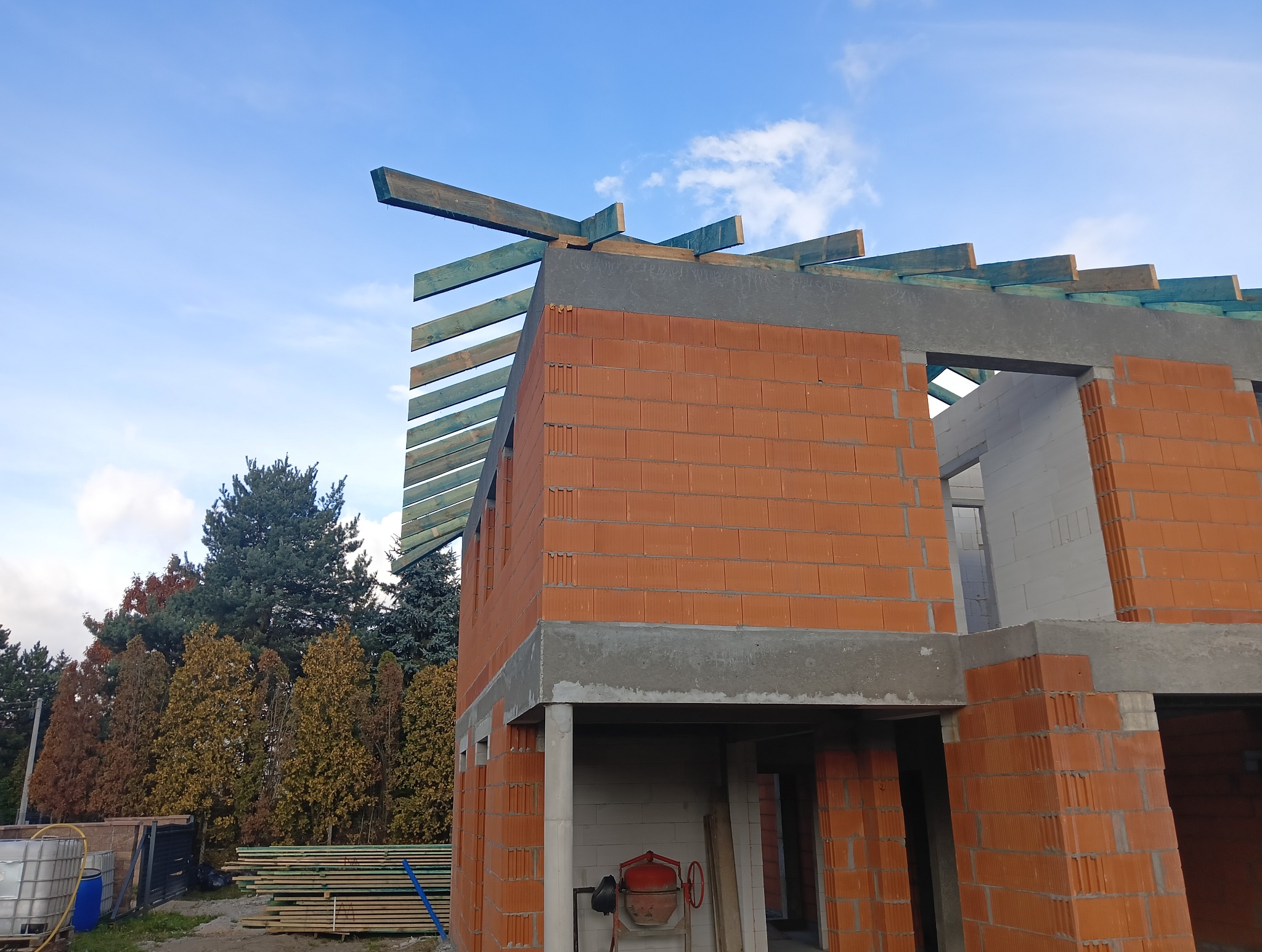 Surowy stan domu murowanego z czerwonej cegły ceramicznej z widocznymi elementami szalunku i drewnianymi belkami więźby dachowej na tle błękitnego nieba z chmurami