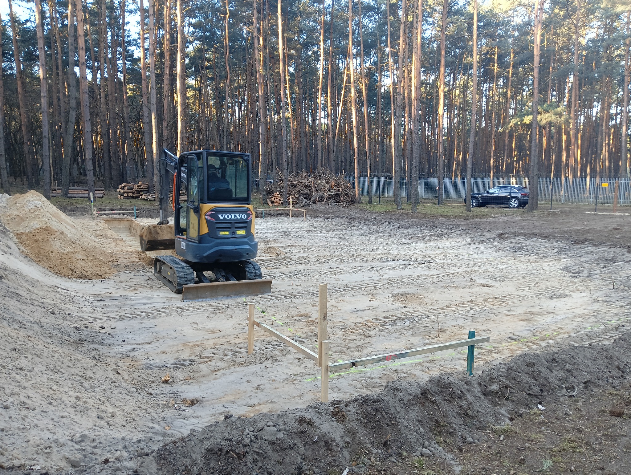 Żółto-szara mini koparka Volvo na placu budowy z wyznaczonymi fundamentami, w tle wysoki las iglasty i zaparkowany czarny samochód.