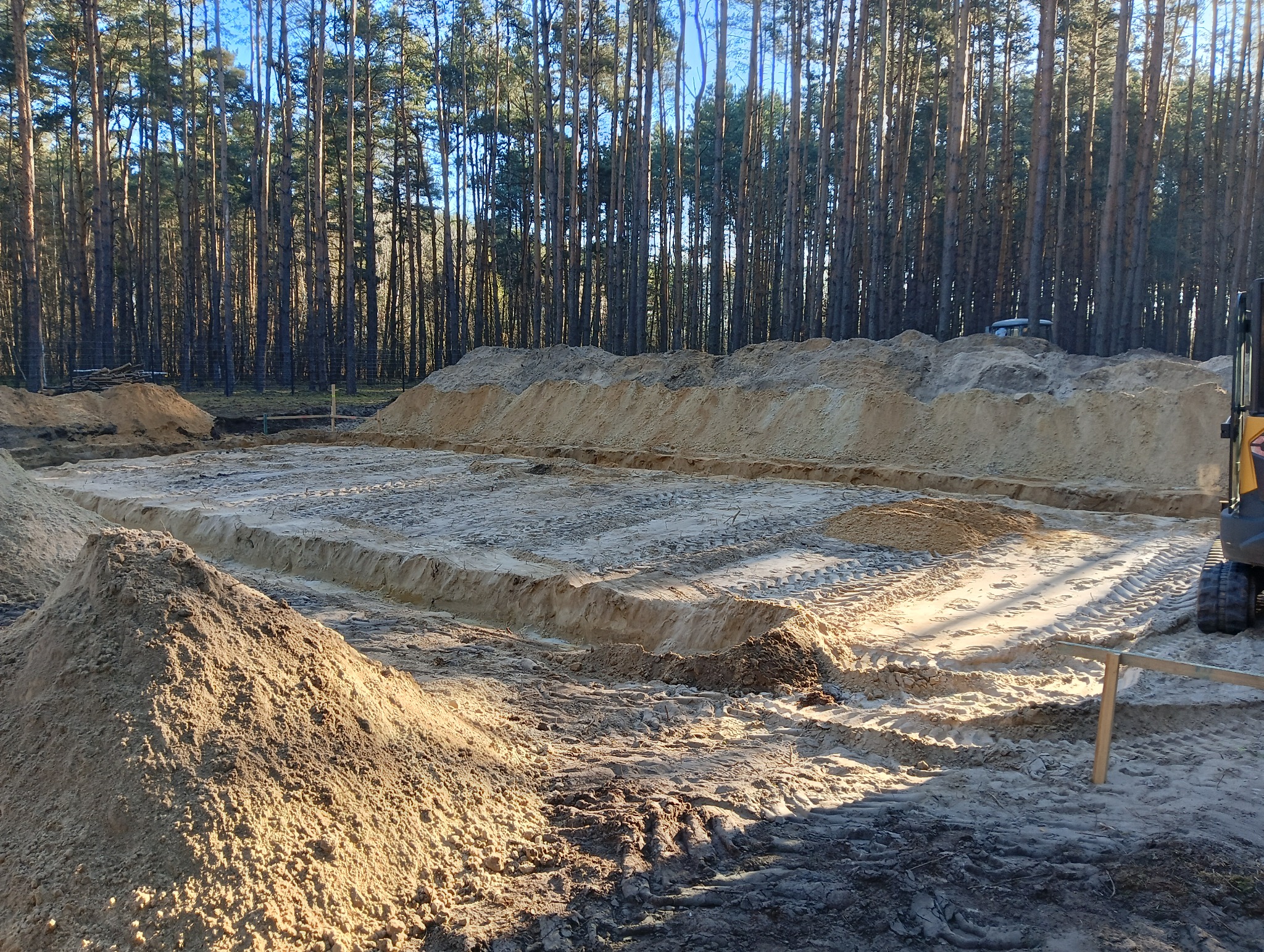 Przygotowany teren pod fundamenty domu z wykopanymi rowami, widoczny stos piasku i fragment koparki na tle lasu sosnowego.