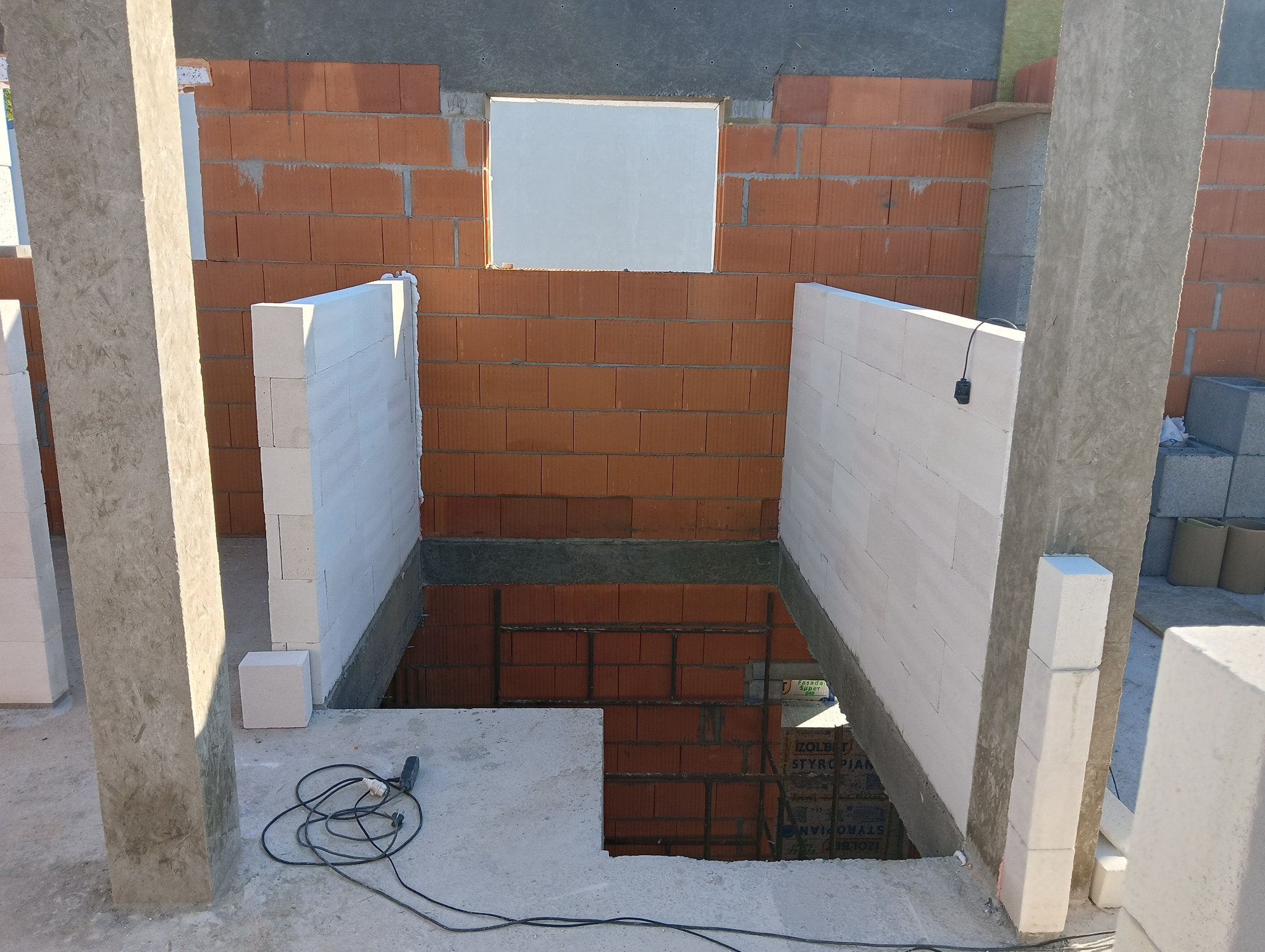 Wznoszenie ścian fundamentowych z bloczków. Widoczne zbrojenie i otwór okienny w murze z czerwonej cegły. Na pierwszym planie narzędzie do cięcia.