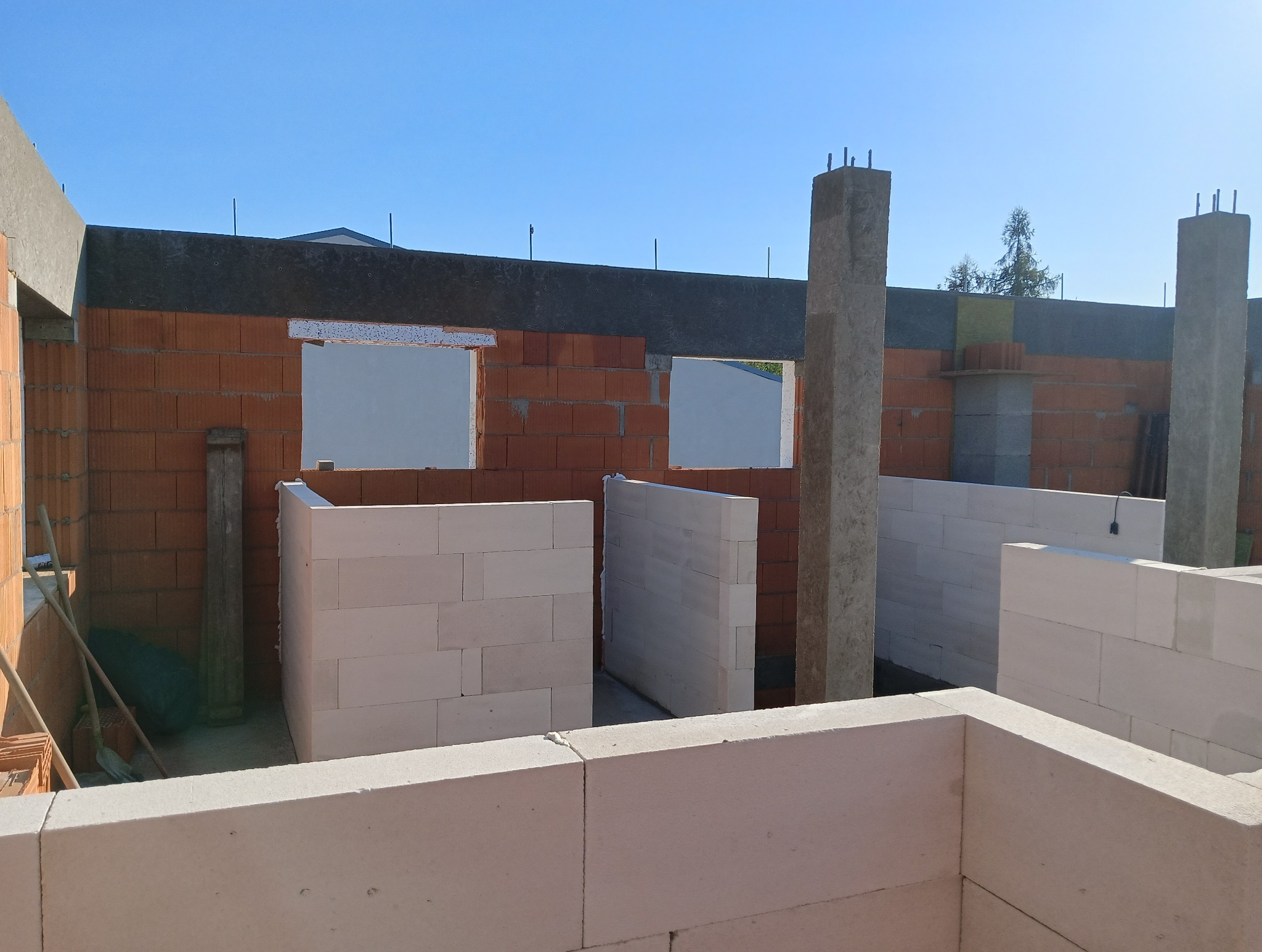 Wznoszenie ścian z bloczków betonu komórkowego Ytong, widoczne zbrojenia pionowe. Ściany z czerwonej cegły, okna w trakcie montażu. Budowa domu w słoneczny dzień.