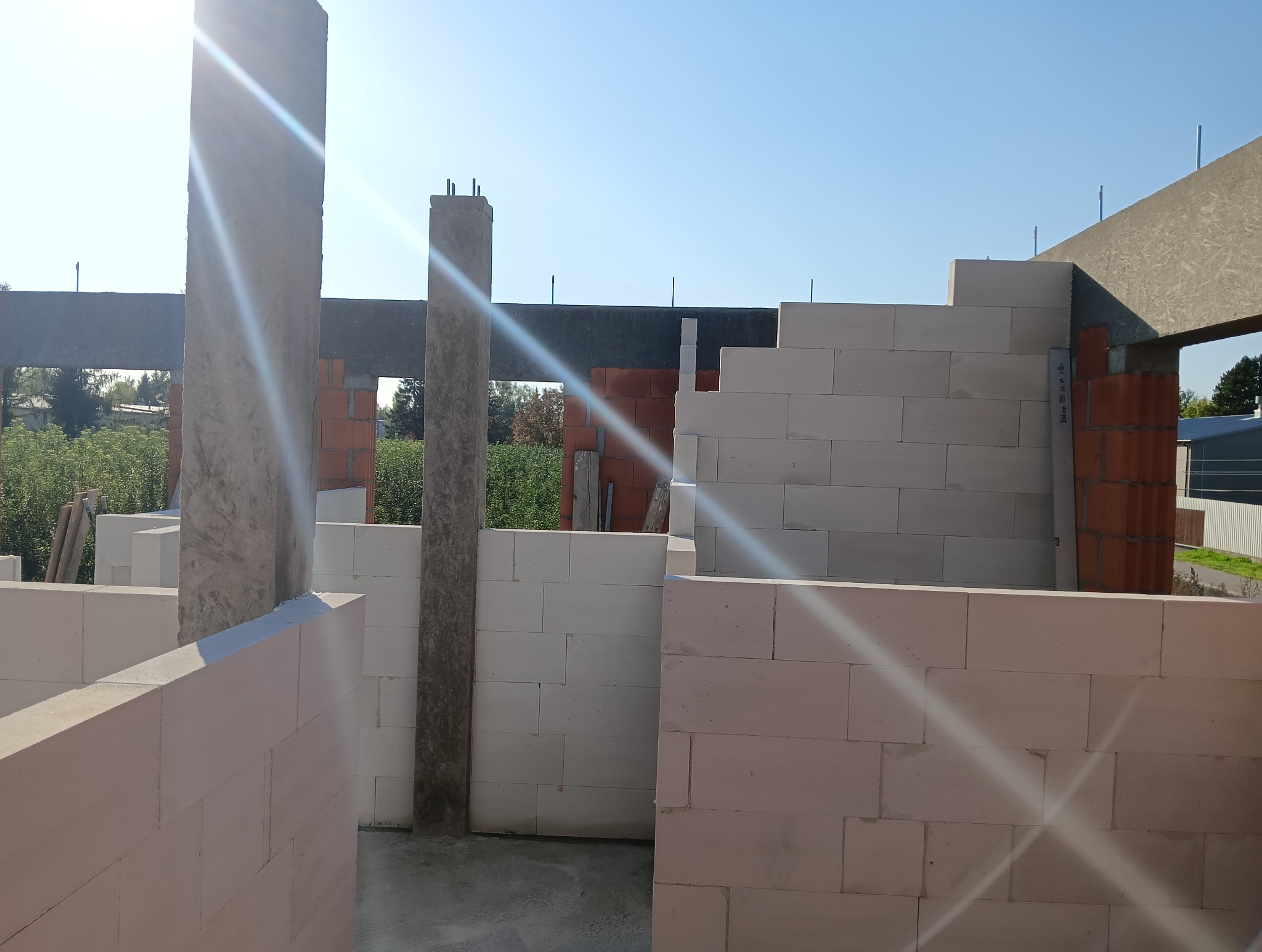 Wznoszenie ścian z bloczków betonu komórkowego. Widoczne zbrojone słupy i nadproża. Budowa w toku, świeże ściany w surowym stanie, w tle zieleń.