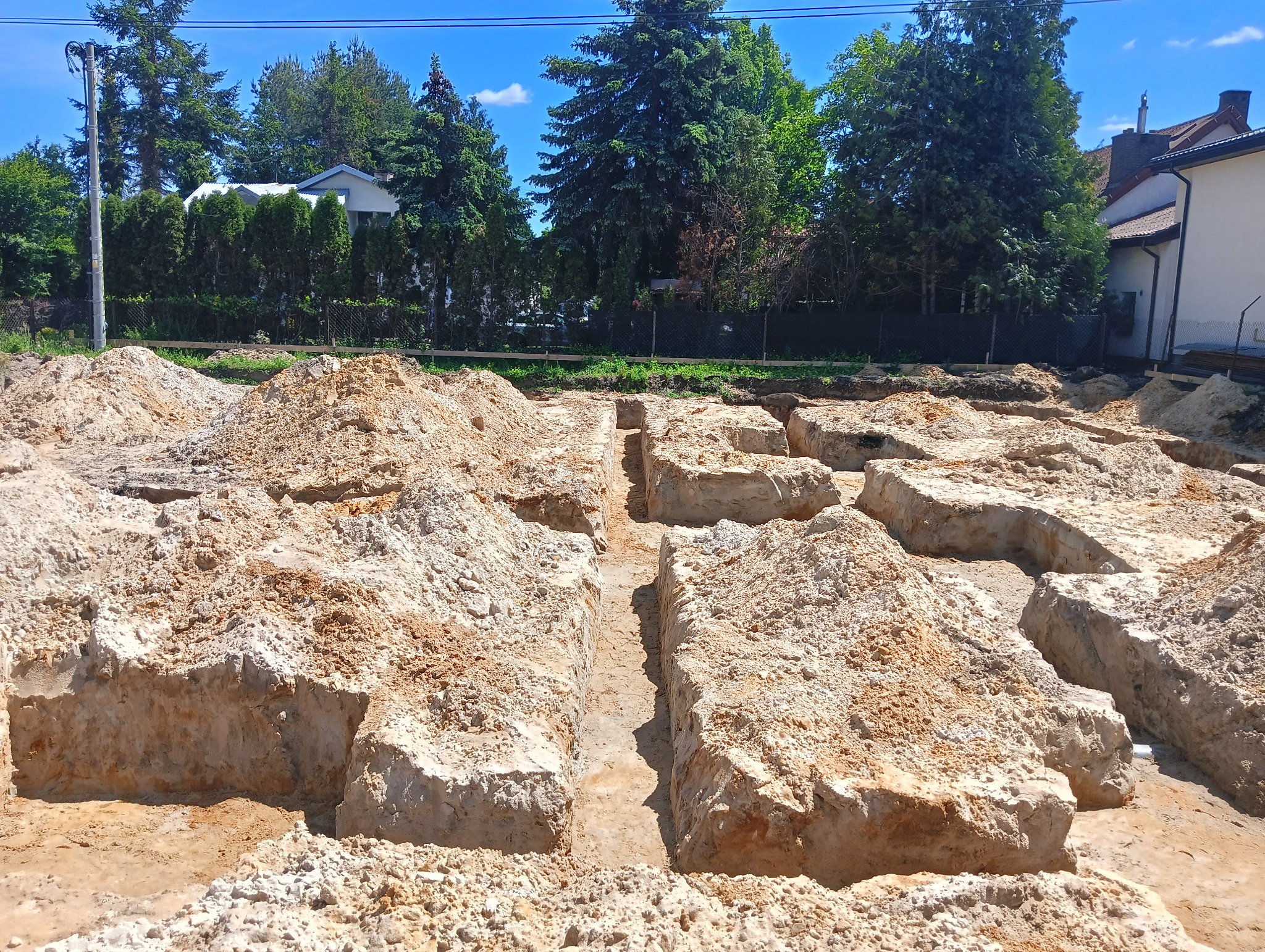 Wykonane wykopy pod fundamenty domu jednorodzinnego. Widoczne wykopane rowy w ziemi, przygotowane do dalszych prac budowlanych. W tle zieleń drzew i budynek.