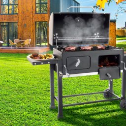 DESIGN M8 - Ogród z nowoczesnym domem i grillem węglowym z otwartą pokrywą, na ruszcie cztery steki, na bocznej półce talerz z grillowanymi ziemniakami i mięsem, widoczny dym i ogień.