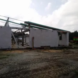 Wznoszenie budynku z bloczków betonowych, widoczne ściany w stanie surowym otwartym, drewniana konstrukcja dachu, drabina oparta o ścianę, w tle wywrotka i hałda piasku.