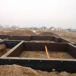 Wykopane i zabezpieczone fundamenty pod budowę domu, widoczne izolacja pionowa, rury kanalizacyjne i otaczający grunt.