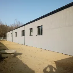 Świeżo otynkowana, szara ściana budynku z kilkoma oknami, widoczny fragment dachu z rynną, piaszczyste podłoże i zarys studzienki kanalizacyjnej z betonową pokrywą.