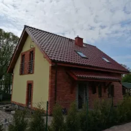 Dwukondygnacyjny dom z czerwonej cegły i otynkowaną elewacją w kolorze jasnej ochry, z dwuspadowym dachem pokrytym czerwoną dachówką ceramiczną, widoczne okna dachowe i komin, w tle drzewa...