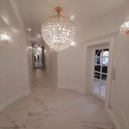 Korytarz w stylu glamour z białymi ścianami, marmurową podłogą i kryształowymi żyrandolami, lustrzana ściana w głębi.