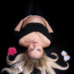 Kobieta w czarnym ubraniu leży na czarnym tle, dłonie na brzuchu ciążowym, blond włosy rozłożone wokół głowy, ozdobiona trzema kwiatami w różnych kolorach.